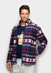 Uomo che indossa una giacca in pile con zip a maniche lunghe rosso, bianco e blu scuro con un motivo, sopra un felpa blu scuro con una grafica di un orsetto in un bicchiere di birra.