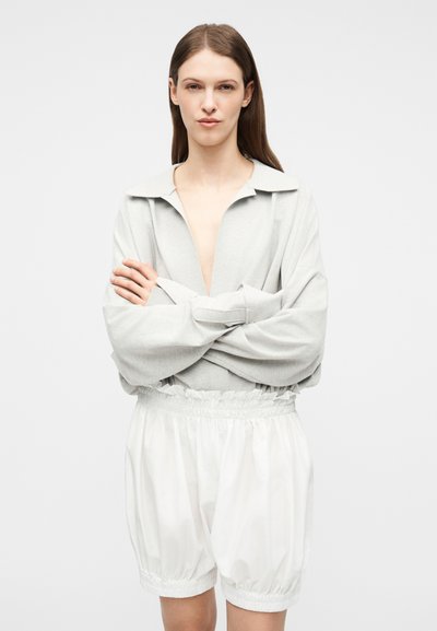 Νεαρή γυναίκα με μακριά καστανά μαλλιά φοράει ανοιχτό γκρι μπλούζα με μακριά μανίκια και βαθύ V-λαιμόκοψη, καθώς και λευκά σορτσάκια με ελαστική μέση, με τα χέρια σταυρωμένα.