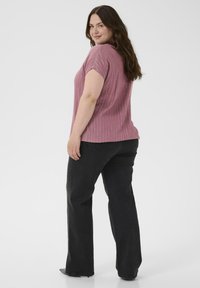 Kvinne med langt brunt hår, iført en rosa stripete kortermet topp og sorte jeans, som står vendt litt til siden og smiler.