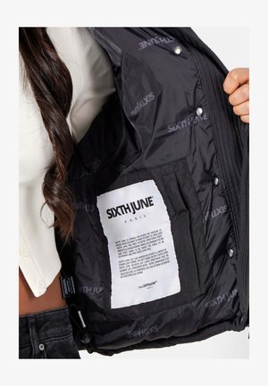 Vrouw die de binnenkant van een zwarte gewatteerde jas laat zien met een groot wit label van het merk SIXTH JUNE Paris en een tekstpatch.