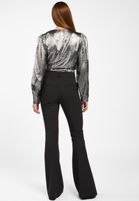 Vrouw met lang rood haar in een glanzende zilveren top met lange mouwen en zwarte hoge taille flare-pantalons, staand met de rug naar de kijker.