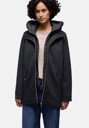 Femme aux cheveux bouclés portant des manteaux noirs zippés superposés sur un haut beige et un jean bleu, debout devant un fond uni.