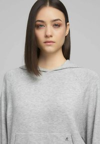 Sudadera con capucha de color gris claro, con cordón regulable, textura suave y bolsillo delantero. Cuenta con mangas tipo raglán y un pequeño logo en la parte inferior izquierda.