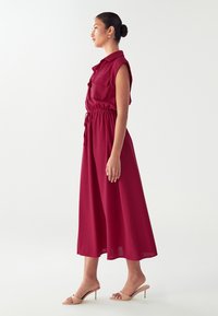 Abito rosso con maniche corte, vita elastica e due tasche. Tessuto morbido con gonna fluida, abbinato a sandali con tacco beige.