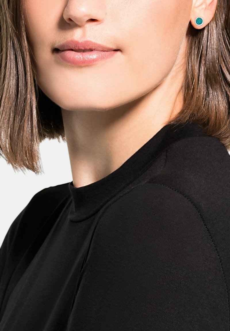 Femme aux cheveux bruns courts portant un top noir et des boucles d'oreilles vertes, lèvres hermétiquement closes et souriant légèrement, sur fond blanc.