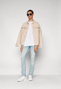 Chaqueta oversized de color beis claro, camiseta blanca, jeans desgastados de color azul claro y zapatillas blancas. Los accesorios incluyen unas gafas de sol negras.