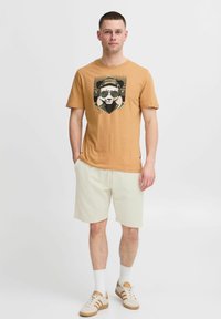 T-shirt à manches courtes, couleur moutarde, avec un graphique de panda en lunettes de soleil, associé à un short clair et des chaussures de sport beiges.