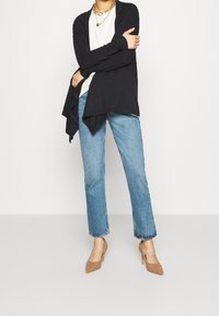 Un persona che indossa un cardigan nero, una maglietta bianca, jeans blu, tacchi alti beige e una collana dorata spessa, in piedi contro uno sfondo chiaro e semplice.