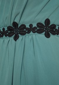 Täckande klänning i teal med rynkad tyg och en svart broderad blommig bälte detalj vid midjan, vilket ger textur och kontrast.