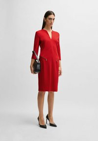 Rotes Kleid mit V-Ausschnitt, drei Viertelärmeln und zwei Reißverschlusstaschen. Kombiniert mit einer schwarzen Handtasche und schwarzen spitzen Pumps.