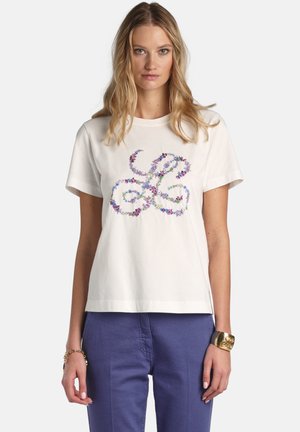 Mujer con una camiseta blanca con un diseño floral en espiral y pantalones azules, complementada con una pulsera de oro y una pulsera de cadena.