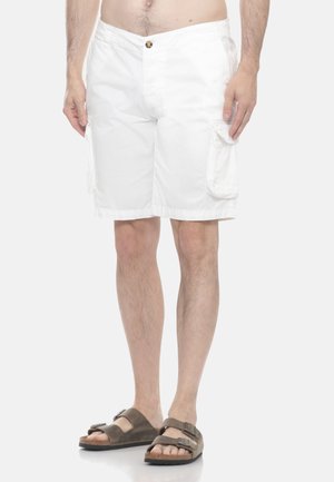 Uomo con pantaloncini cargo bianchi con tasche laterali e sandali marroni a doppio cinturino, in piedi davanti a uno sfondo chiaro e uniforme.
