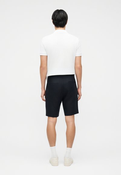 Άνδρας που στέκεται με την πλάτη γυρισμένη, φοράει λευκό κοντομάνικο πόλο, μαύρη βερμούδα, λευκές κάλτσες και μπεζ αθλητικά παπούτσια σε απλό φόντο.