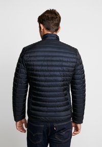 Marinblå puffjacka med quiltad design, hög krage och dragkedjefickor. Tillverkad av glänsande, lätt material. Figursydd passform, texturerad yta.