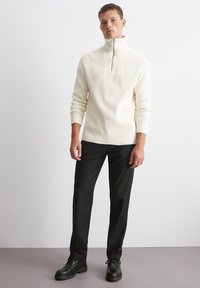 Cremfarbener, gerippter Pullover mit einem halben Reißverschluss am Kragen, kombiniert mit schwarzen gestreiften Hosen und schwarzen Schnürschuhen. Flache Textur, lässige Passform.