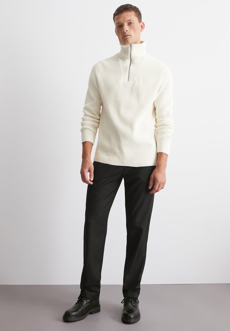 Cremfarbener, gerippter Pullover mit einem halben Reißverschluss am Kragen, kombiniert mit schwarzen gestreiften Hosen und schwarzen Schnürschuhen. Flache Textur, lässige Passform.