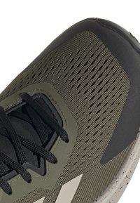 Sportskor med ett mörk olivgrönt textilöverdrag, svarta accenter, perforerade sektioner för andningsförmåga och en fläckig grå sula.