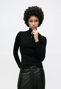 Schwarzer, gerippter Rollkragenpullover mit langen Ärmeln und goldenen Knopfakzenten auf der Schulter, kombiniert mit schwarzen, hoch taillierten Lederhosen.