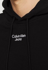 Černá mikina s kapucí se zaoblenou kapsou na přední straně, stahovacími šňůrkami a bílým vyšitým logem "Calvin Klein Jeans" na hrudi. Měkká látka.