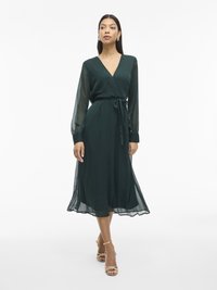 Sötétzöld, térdígy érő, könnyű szövetből készült bodycon ruha. Hosszú áttetsző ujjaival, V-kivágásával és megkötős derékkal rendelkezik. A fodros szegély kiemeli a megjelenését.