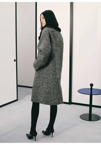 Cappotto grigio in tweed con vestibilità ampia, tasche laterali e colletto alto. Indossato sopra collant neri e tacchi. Sfondo minimalista.