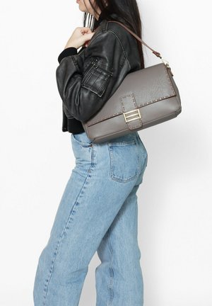 Borsa grigia con texture, dotata di una chiusura color oro e cuciture marroni, indossata a spalla, insieme a una giacca nera e jeans in denim chiaro.