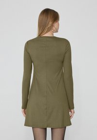 Vestito a maniche lunghe a coste in verde oliva, caratterizzato da una linea dritta con una cucitura sul retro. Lunghezza al ginocchio con un tessuto testurizzato.