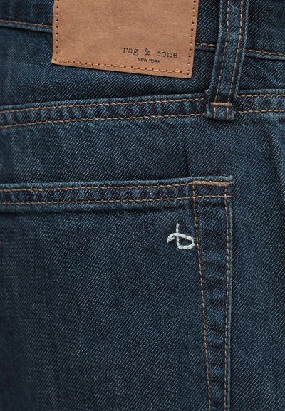 Donkerdenim jeans hebben een bruine leren label met de tekst "rag & bone NEW YORK". Een witte stiksel detail is zichtbaar op de achterzak.