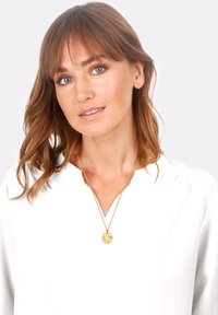 Collana con pendente d'oro su una catena sottile, con un ciondolo rotondo. Il modello indossa una camicia bianca con scollatura a V. I capelli sono lunghi fino alle spalle.