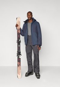 Die Skiausrüstung besteht aus einer marineblauen wasserdichten Jacke, einem grauen Langarmshirt, grauen Hosen und schwarzen Skistiefeln. Hält ein Paar Skier.