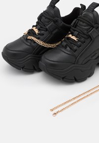Black chunky sneakers made of leather with a thick sole, featuring a gold chain accent across the laces, alongside a matching gold chain.
