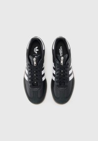 Pár černých tenisek Adidas s bílými pruhy a černými tkaničkami, pohled shora na jednobarevném světlém pozadí.