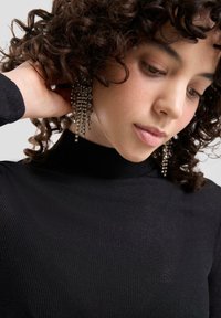 Czarny prążkowany golf. Modelka o kręconych włosach nosząca długie srebrne kolczyki z kryształkami. Skupienie na teksturze tkaniny i szczegółach kolczyków.