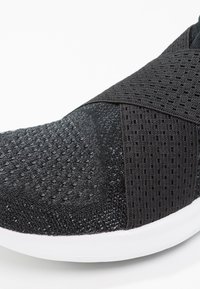Chaussure de sport noire présentant un dessus en tricot texturé avec des détails en mesh, une sangle élastique au niveau du métatarse et une semelle en caoutchouc blanche.
