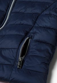 Veste matelassée bleu marine avec un design à motifs piqués, dotée d'une poche zippée argentée et d'une texture lisse. Les accents comprennent un tirette de fermeture éclair métallique.