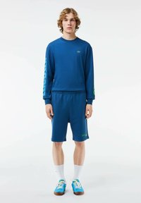 Sudadera de manga larga azul y pantalones cortos a juego en tela suave. Presenta un logo en el pecho y un acento verde vibrante en los pantalones cortos. Medias blancas y zapatos azules.