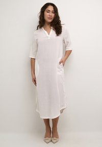 Robe en lin blanc avec un décolleté en V, des manches trois-quarts et deux poches latérales. Présente une coupe décontractée et un ourlet légèrement arrondi.