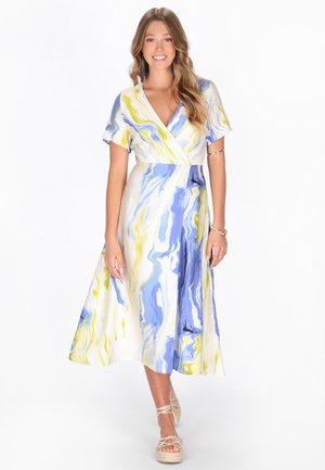 Jeune femme souriante, portant une robe midi à manches courtes avec des motifs tourbillonnants bleu, blanc et jaune, et des sandales beiges à enfiler.