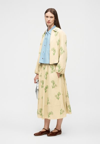 Γυναίκα που φοράει μπεζ λουλουδάτο σακάκι και φούστα με μπλε πουκάμισο με κουμπιά, κρατώντας ασημένια τσάντα και φορώντας καφέ παπούτσια Mary Jane.