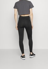 Zwarte legging met een hoge tailleband, een gladde textuur en een logo-accent op de achterkant. Gepaard met een cropped grijs t-shirt en zwarte sneakers.