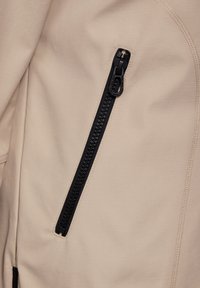 Tissu de manteau beige avec une texture lisse, comportant une poche zippée noire bien visible avec un détail décoratif sur la fermeture éclair.