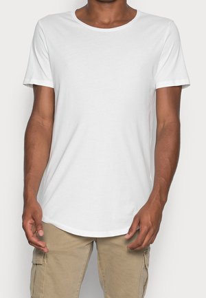 Hombre viste una camiseta blanca de manga corta y pantalones cargo beige, de pie con los brazos relajados a los lados sobre un fondo claro.