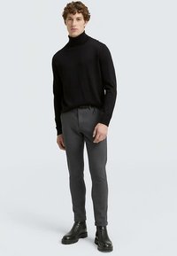 Črni pulover z visokim ovratnikom v oprijeti zasnovi, kombiniran z sivimi hlačami ozkega kroja. Model nosi črne gležnjarje. Enostavno ozadje.