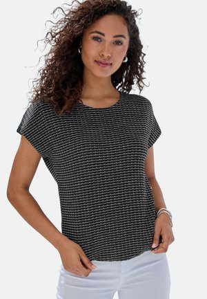 Frau mit lockigem Haar, die ein schwarzes kurzärmeliges Oberteil mit weißen horizontalen Wellenmustern und weiße Hose trägt, posiert mit den Händen nahe der Taille.