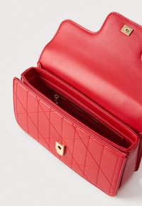 Crvena kožna torba s prošivenim uzorkom, zlatnim kopčama i unutarnjim džepom na patentni zatvarač. Ima preklopno zatvaranje sa četvrtastim kopčom.