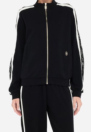 Czarna kurtka na suwak z białymi paskami na ramionach i rękawach, czarne spodnie z białymi paskami po bokach, noszona przez osobę od szyi do bioder.