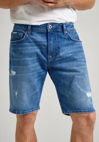 Pantalones cortos de mezclilla, azul medio con parches desgastados, cinco bolsillos, cierre de cremallera y botón. Presenta costuras contrastantes y dobladillo vuelto.