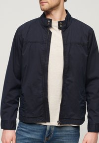 Lekka kurtka bomber w kolorze granatowym, z kołnierzem stojącym, zatrzaskami i przednim zamkiem błyskawicznym. Charakteryzuje się szczegółami przeszyć i elastycznymi mankietami.
