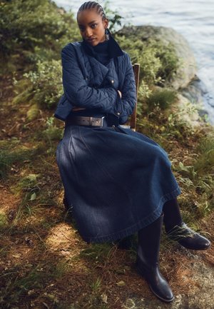 Mujer con el pelo trenzado sentada al aire libre en una silla, con una chaqueta acolchada, falda vaquera, botas negras y pendientes grandes cerca del agua y la vegetación.