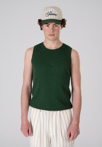 Green ribbed tank top with a round neck, paired with beige pinstriped pants and a white cap with green brim and navy lettering.